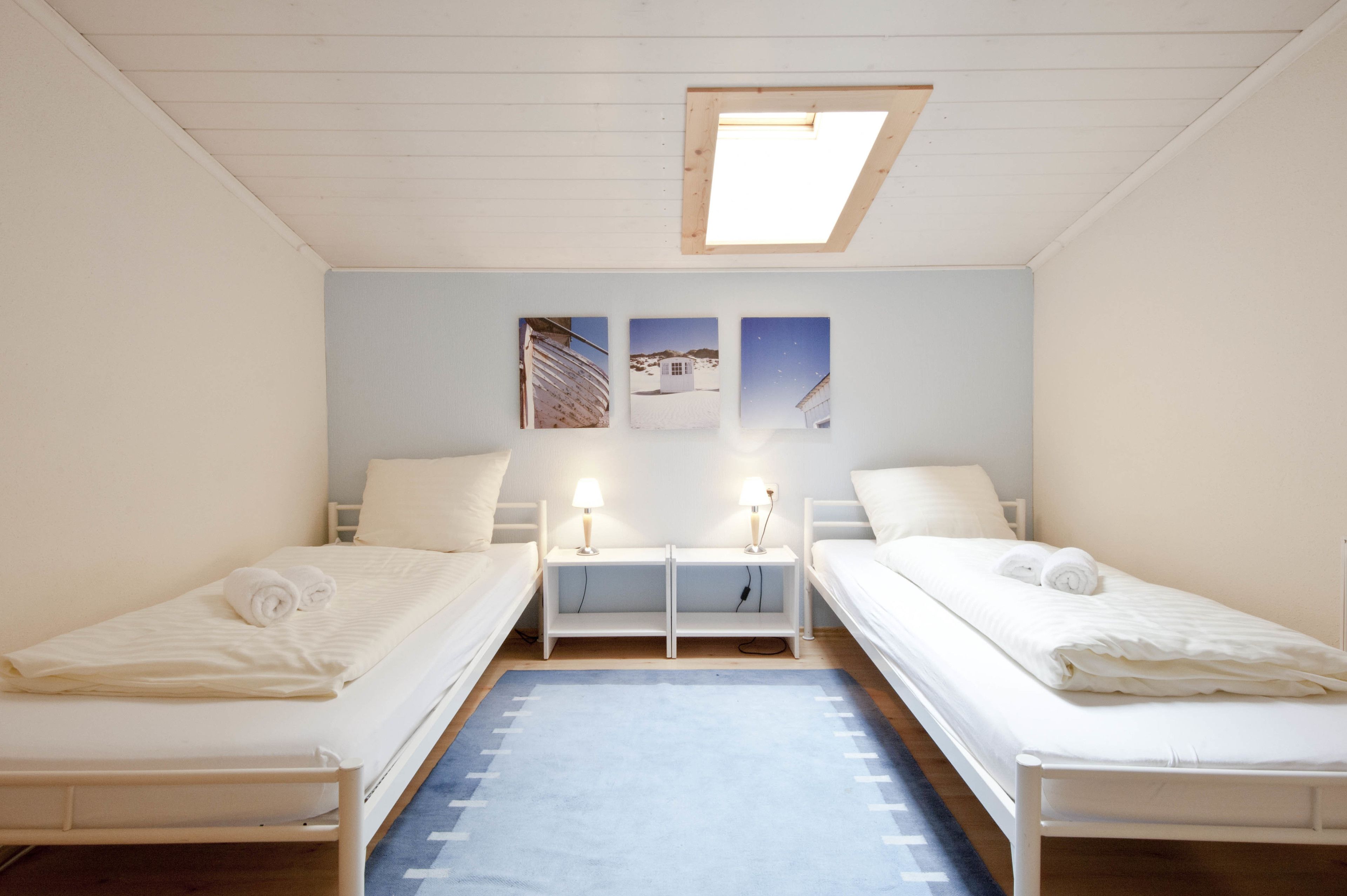 Una habitación con dos camas individuales, un tragaluz, una pequeña mesa con dos lámparas y tres cuadros enmarcados en la pared.