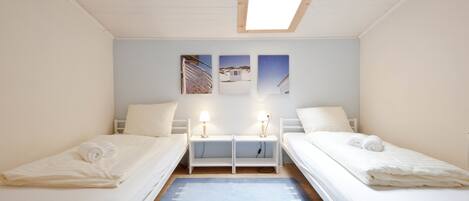 Una habitación con dos camas individuales, un tragaluz, una pequeña mesa con dos lámparas y tres cuadros enmarcados en la pared.
