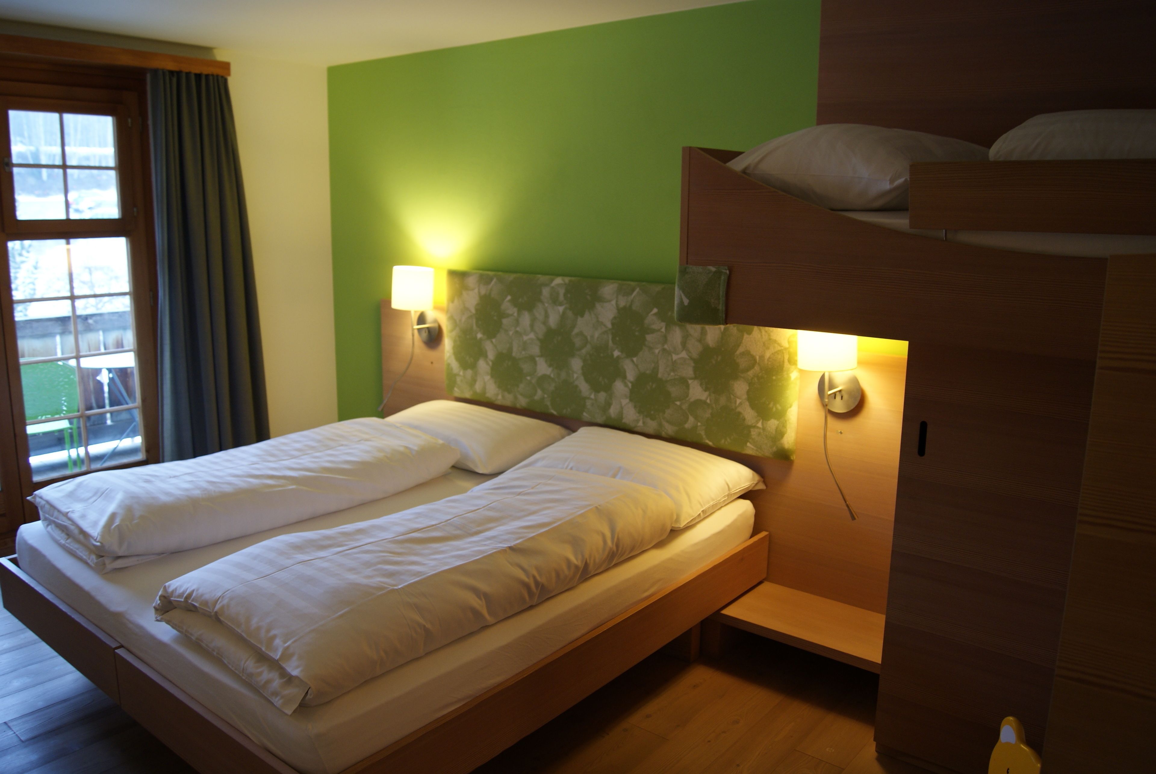A double bed with two bedside tables, a window with curtains, and a bunk bed to the right.