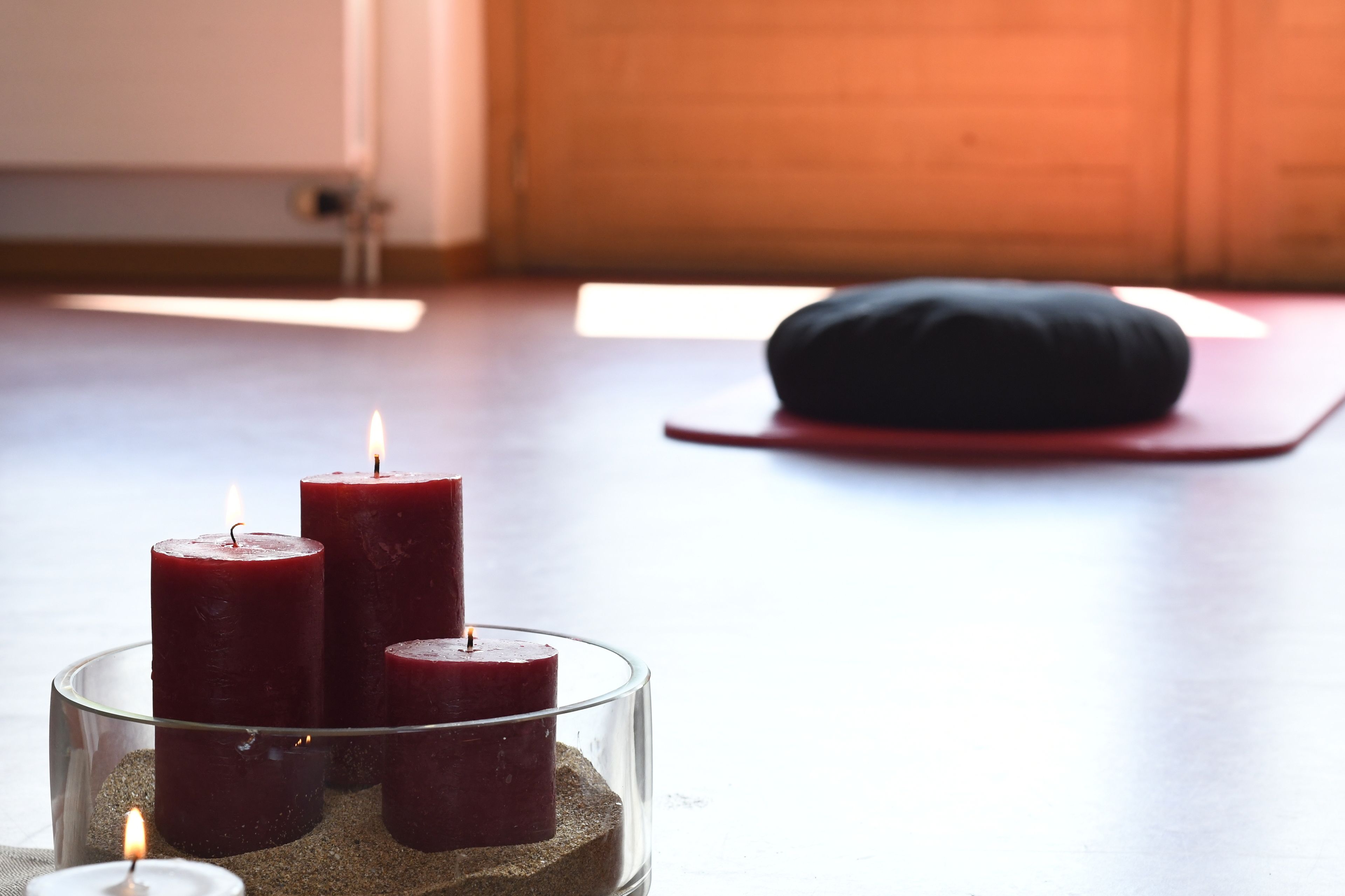 Drei brennende Kerzen in einem Glasgefäß auf Holzboden, mit Yogamatte und Kissen im Hintergrund.