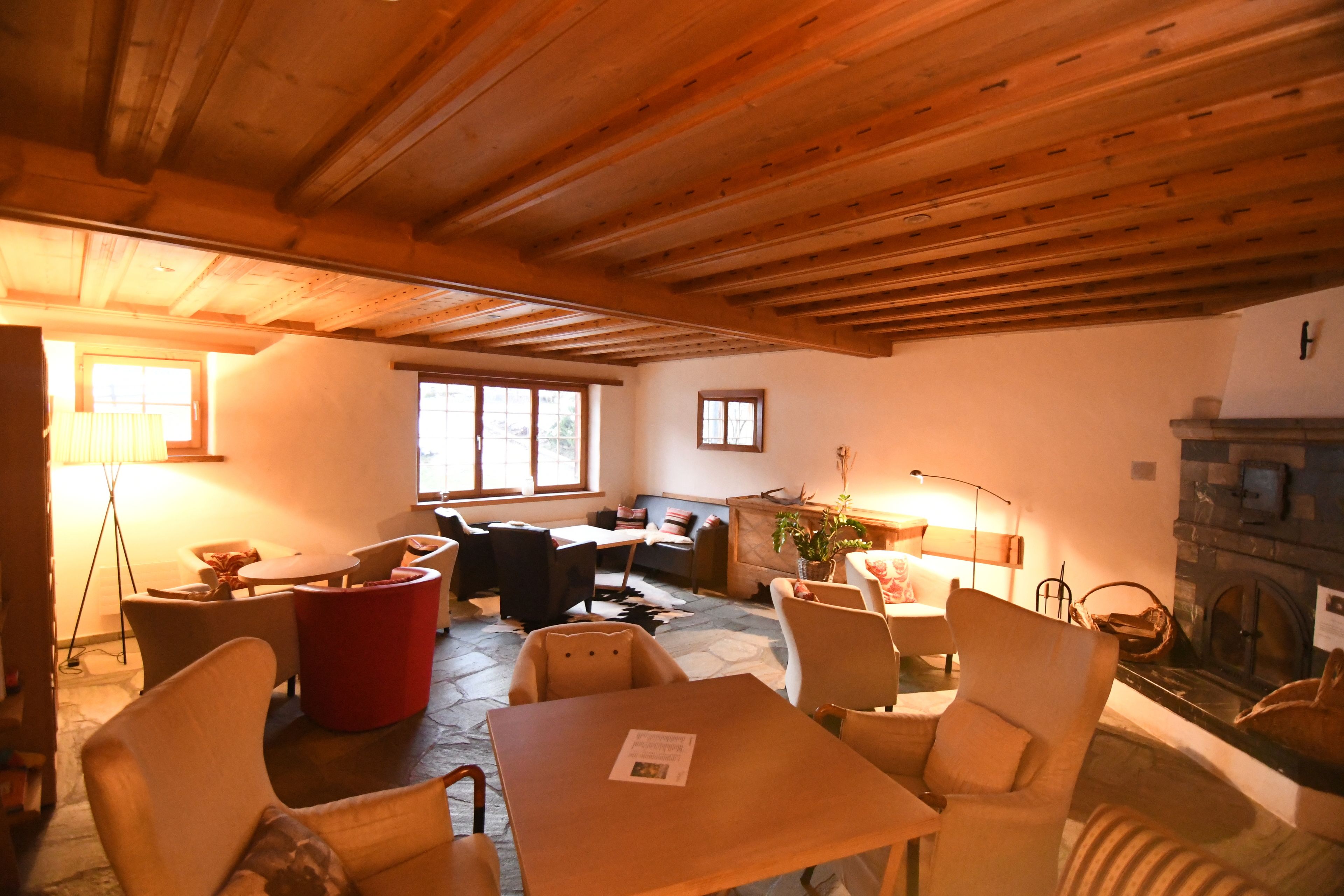 Una habitación acogedora con suelo de piedra, vigas de madera en el techo, una chimenea y una zona de estar con una mesa y sillas.