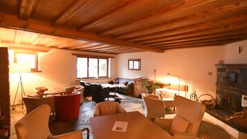 Una habitación acogedora con suelo de piedra, vigas de madera en el techo, una chimenea y una zona de estar con una mesa y sillas.