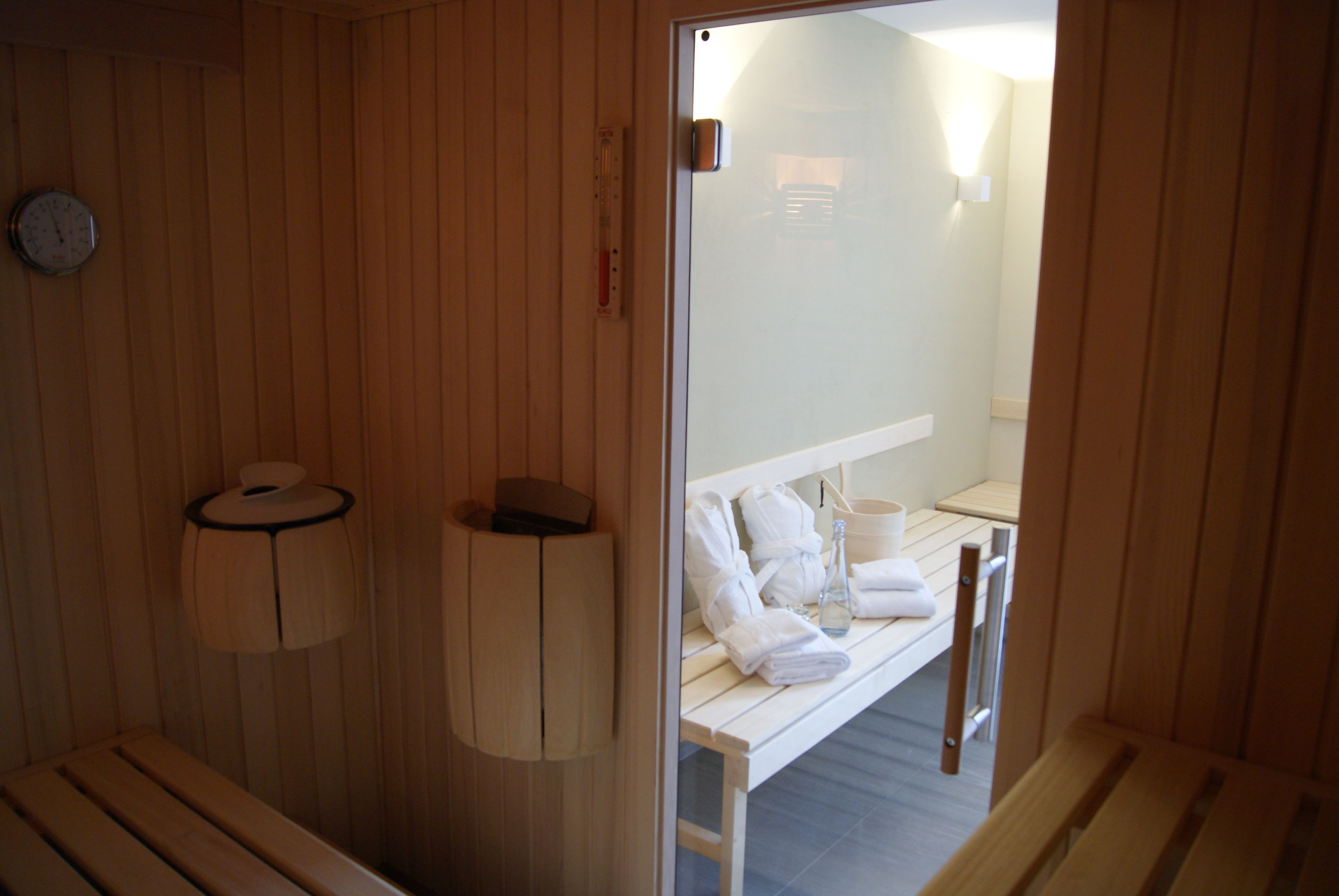 Una sauna de madera con bancos, un perchero para toallas y una puerta de vidrio que da a un baño con lavabo y toallero.