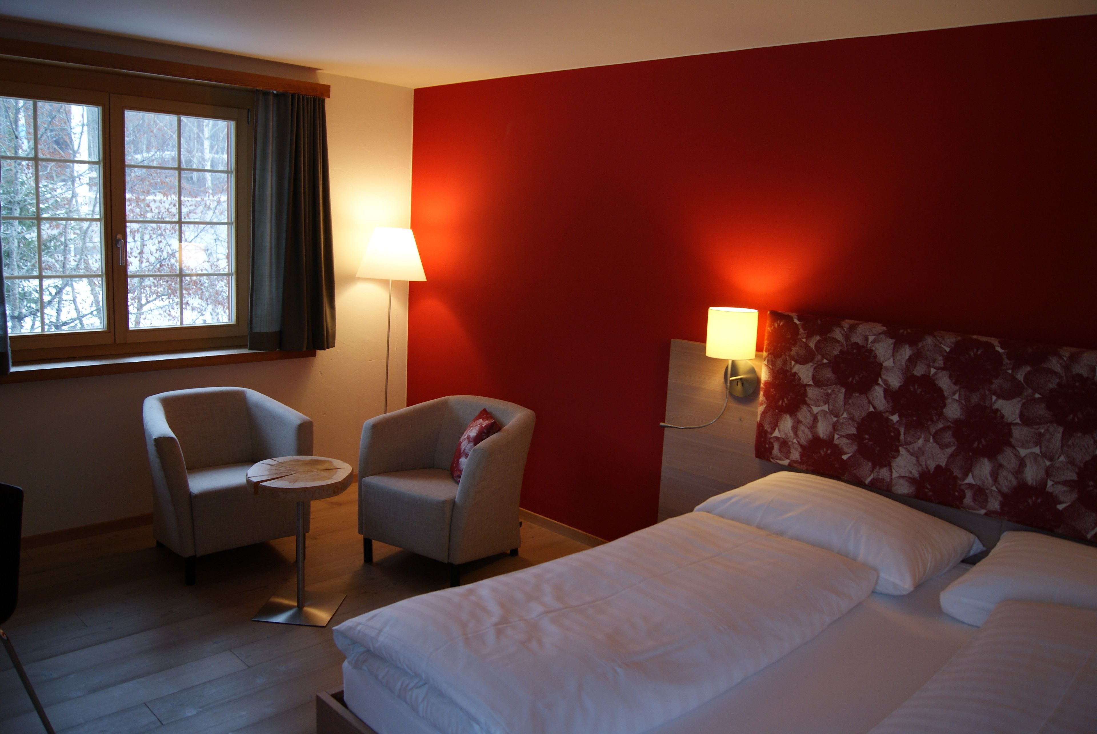 A hotel room with a bed, two armchairs, a small table, a window with curtains, and a red accent wall.