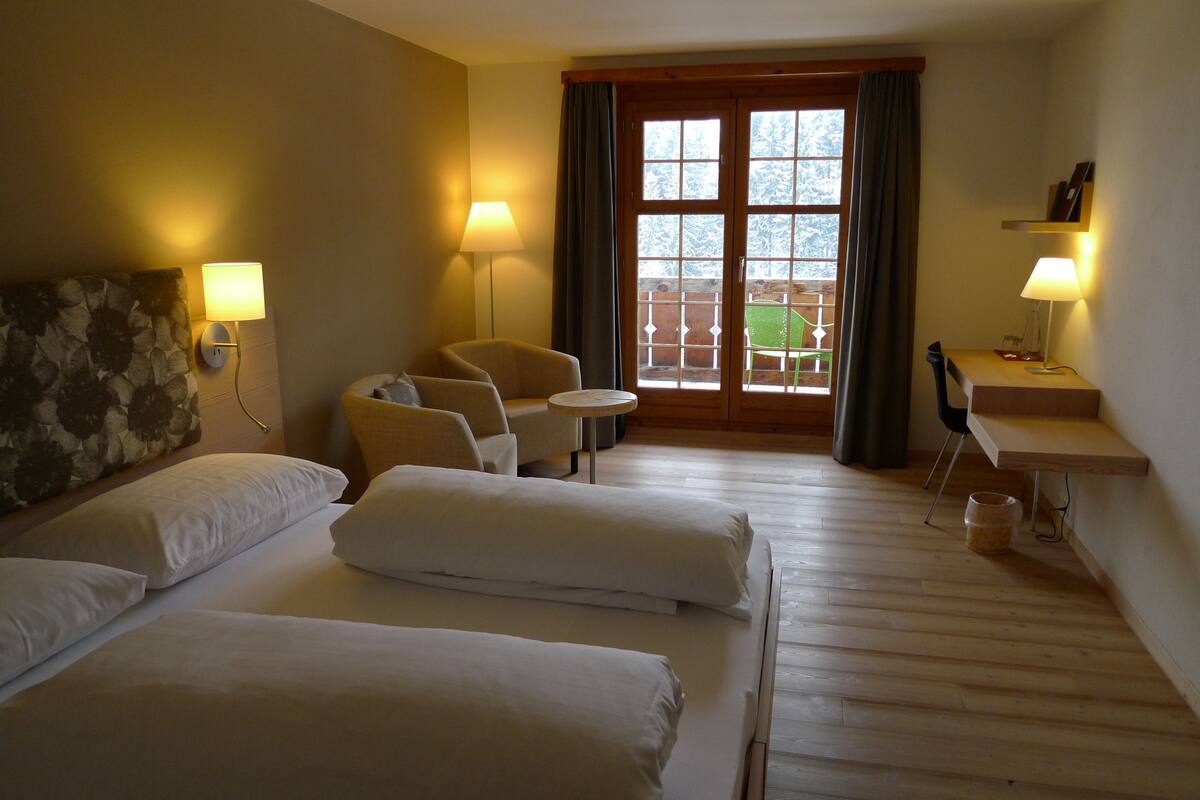 A hotel room with two beds, a desk, a chair, and a small table. There is a window with curtains and a view of a balcony.