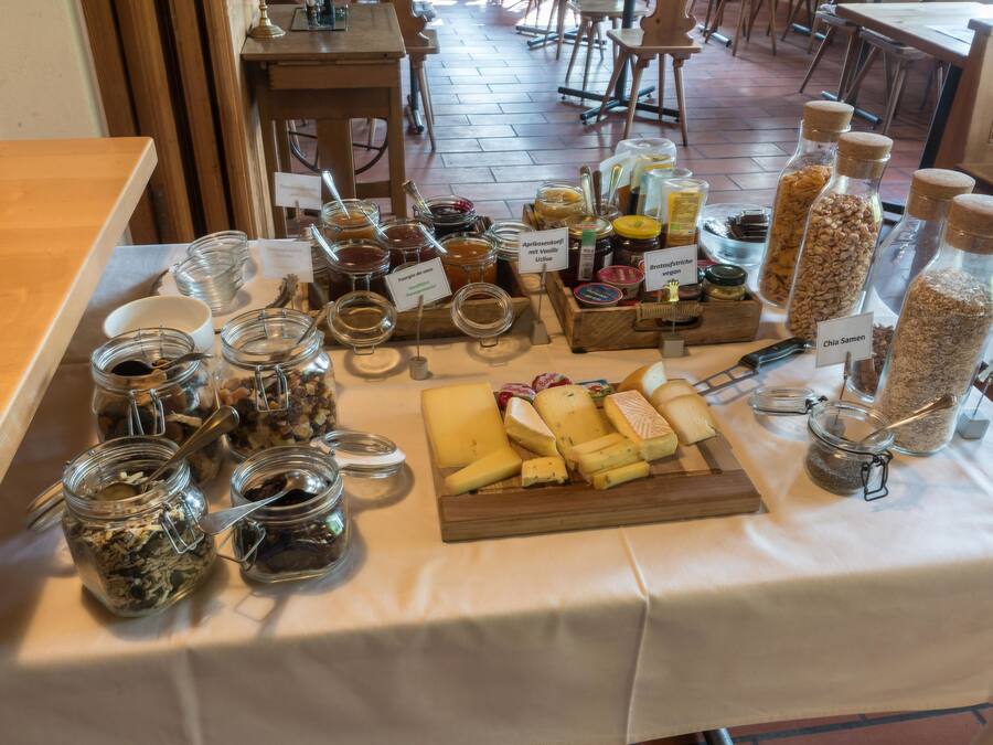 Ein Tisch mit verschiedenen Speisen und Beilagen, darunter Gläser mit Marmelade, ein Holzbrett mit Käse und beschriftete Behälter.