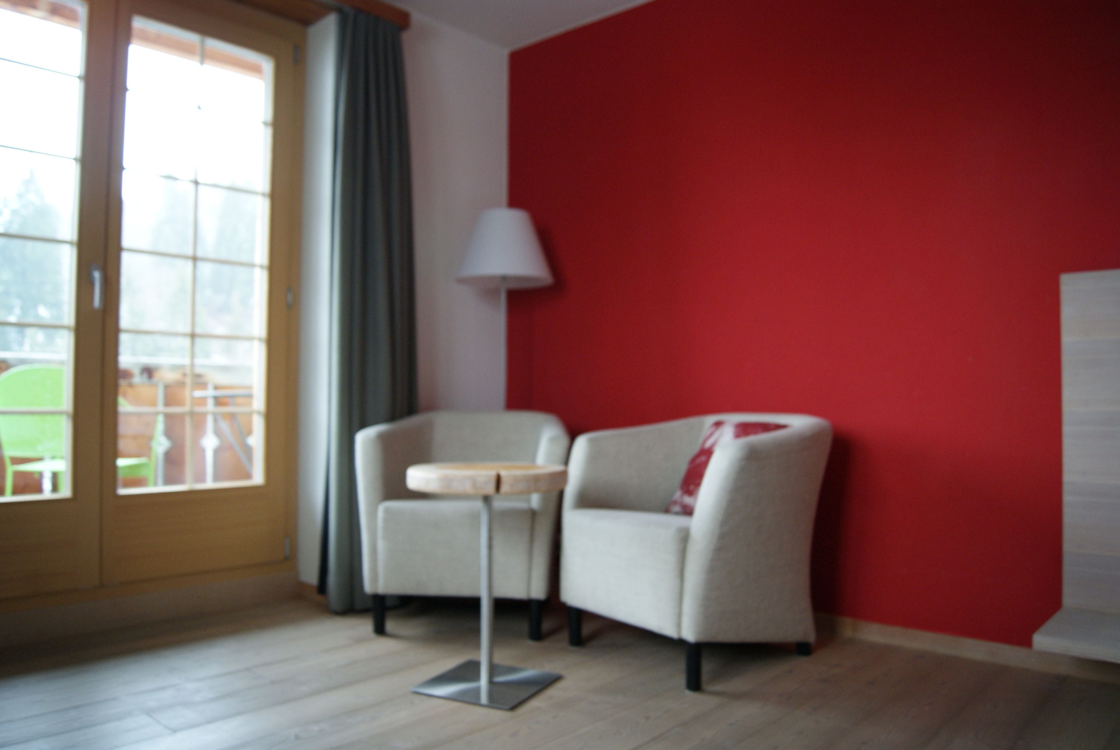 Une pièce avec un mur rouge, une porte à panneaux de verre, un fauteuil beige, une petite table ronde et un lampadaire.