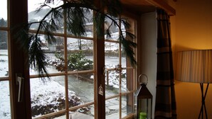 Ein Zimmer mit großem Fenster mit Blick auf eine verschneite Landschaft, ein kleiner Tisch mit einer Laterne und eine Stehlampe.