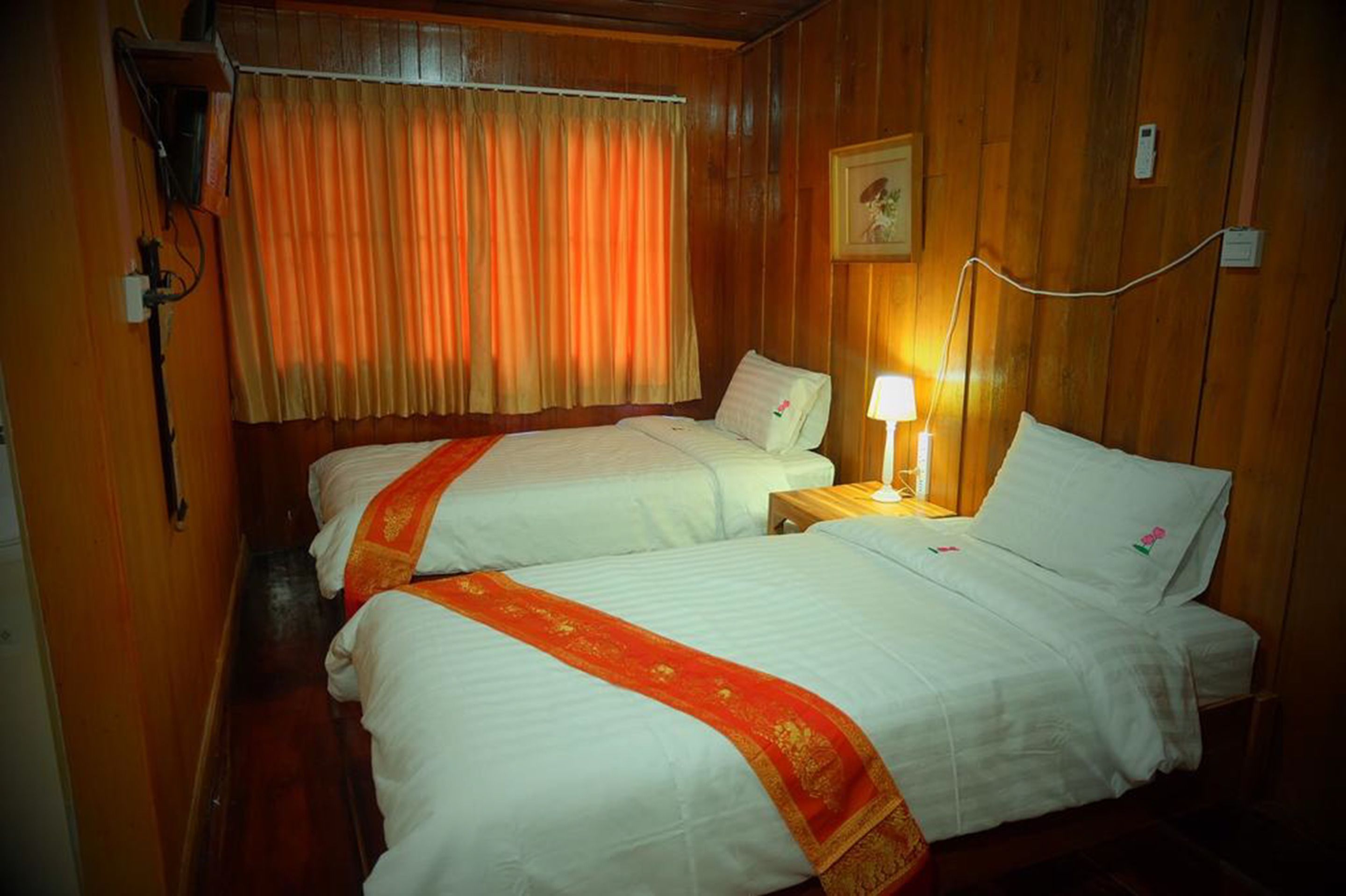 A hotel room with two beds, wooden walls, a window with curtains, a bedside lamp, and a wall-mounted phone.