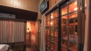 A hotel corridor with wooden walls, a bed, a TV, and large windows with a view of parked cars outside. - The History Cafe' & Guesthouse (Sukhothai)