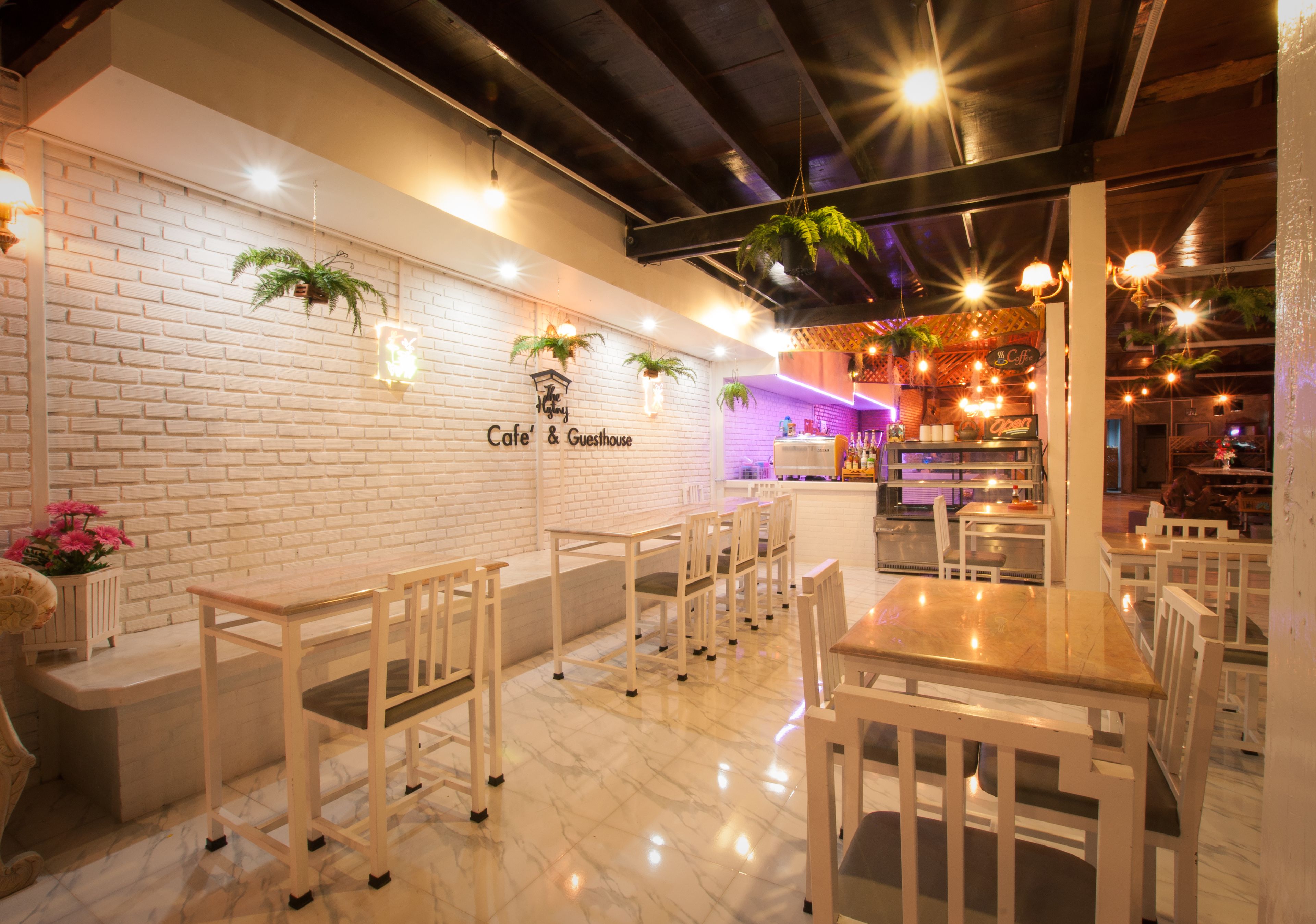 Interior de café e pousada com paredes de tijolos brancos, plantas penduradas e chão de mármore. Mesas e cadeiras dispostas para acomodar visitantes.