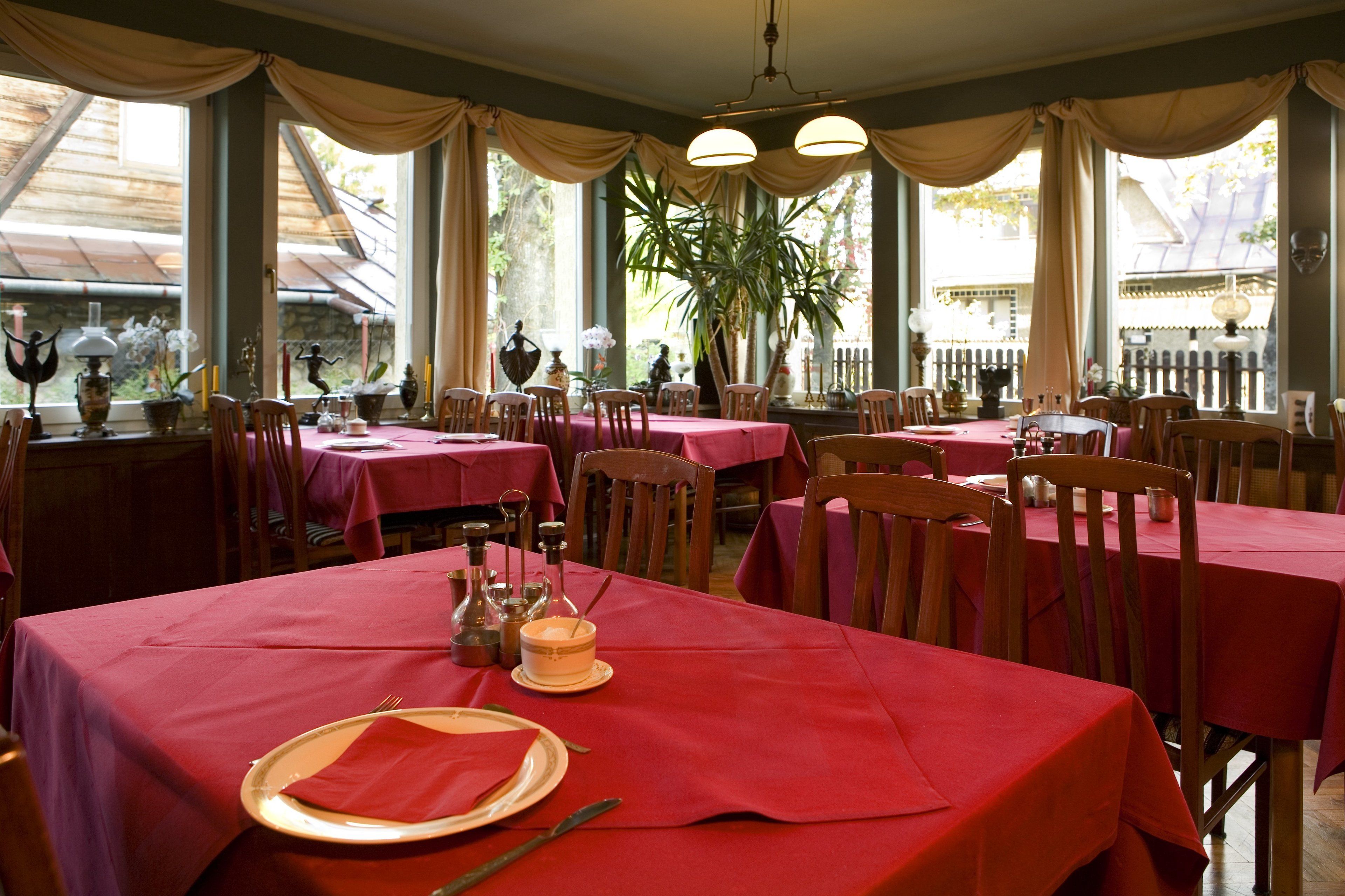 A restaurant with multiple tables covered in red tablecloths, wooden chairs, and a variety of table settings including plates, cutlery, and candles.