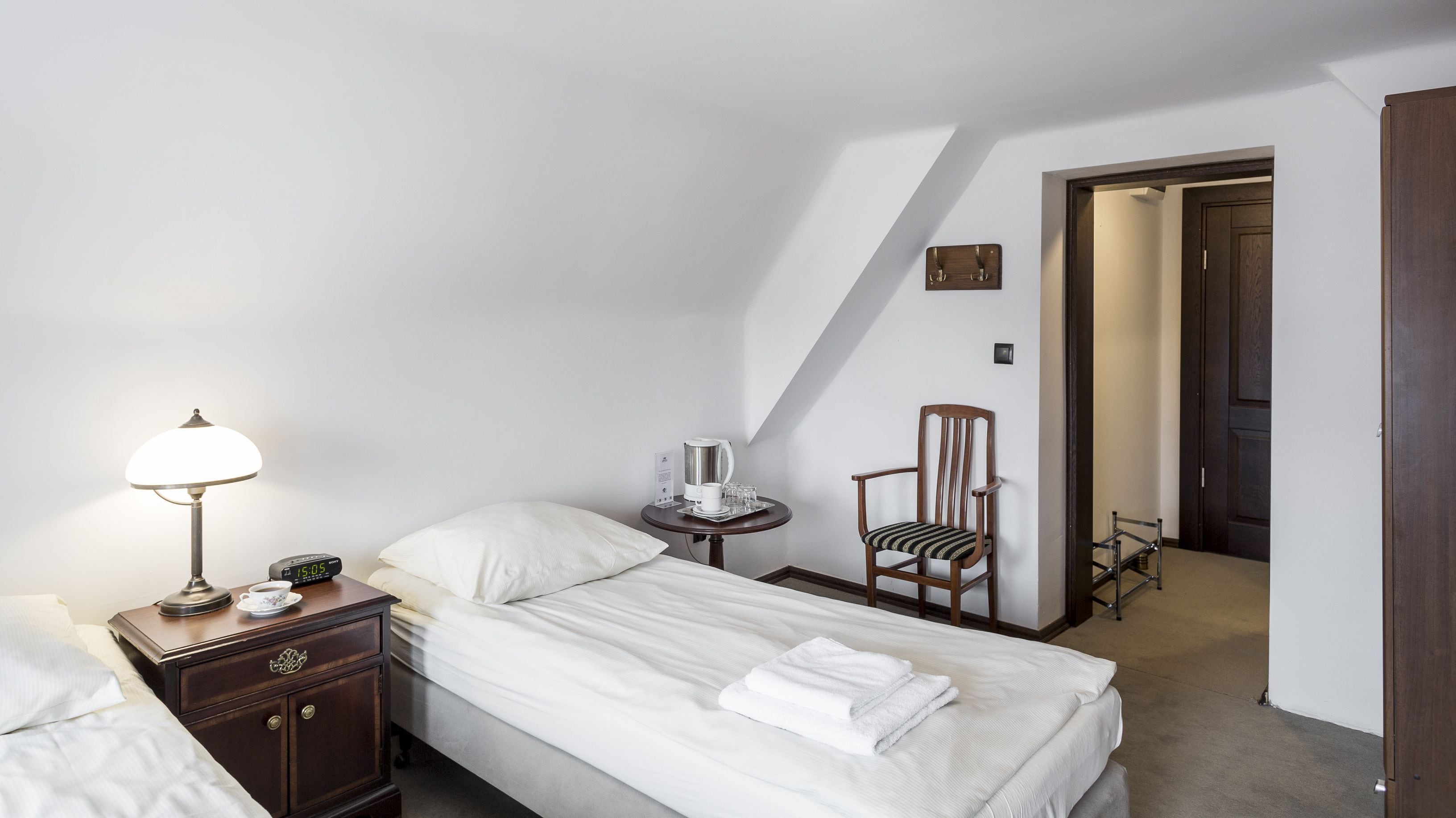 A hotel room with a bed, a bedside table with a lamp, a chair, and a small table with a coffee maker and glasses.