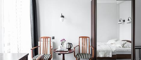 Habitación de hotel con cama, un escritorio y silla, una mesita y otra silla, una lámpara de pared y un espejo que refleja la cama y las luces.