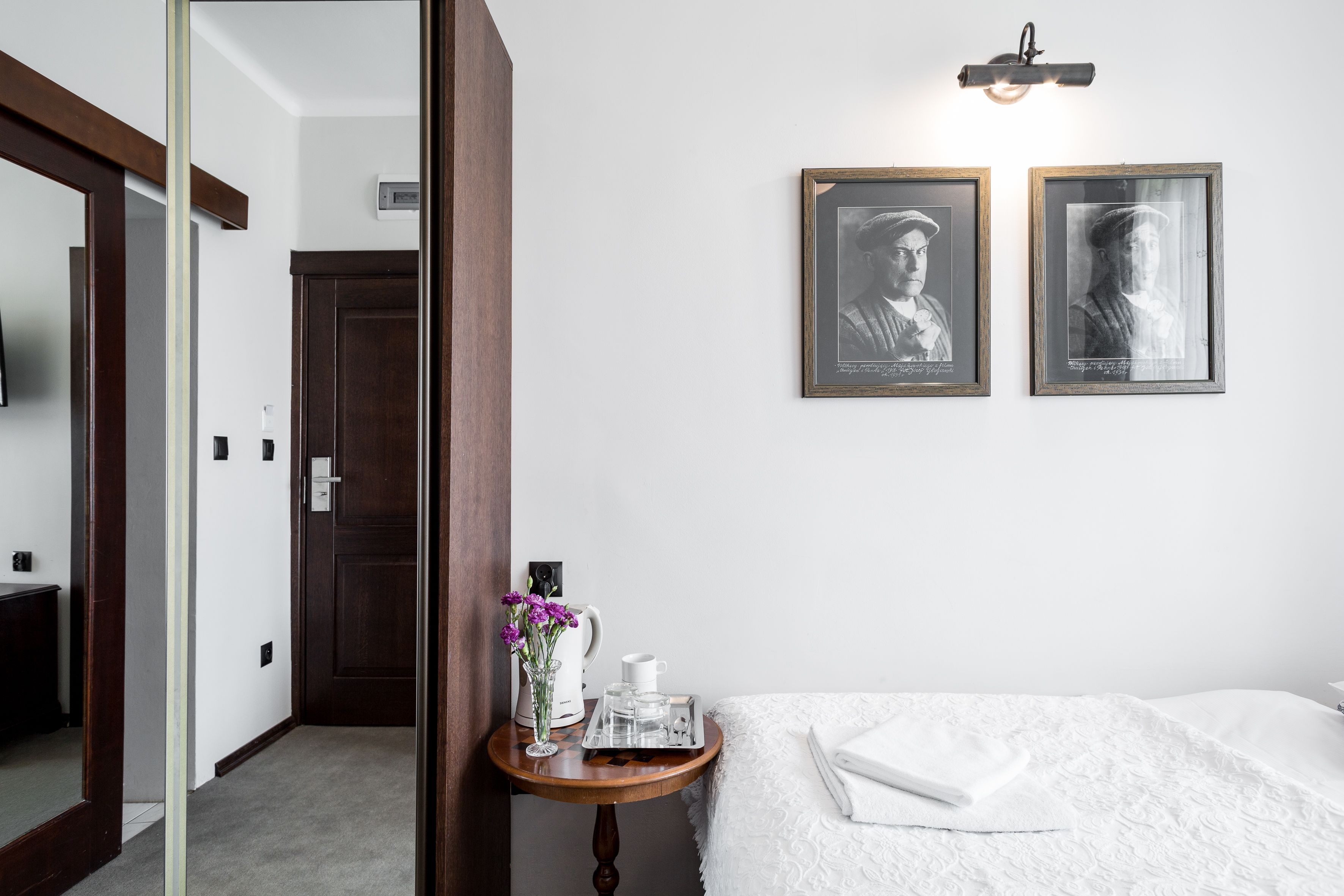 A hotel room with a bed, a bedside table with a vase of flowers, a tray with a cup and a pitcher, and two framed pictures on the wall.