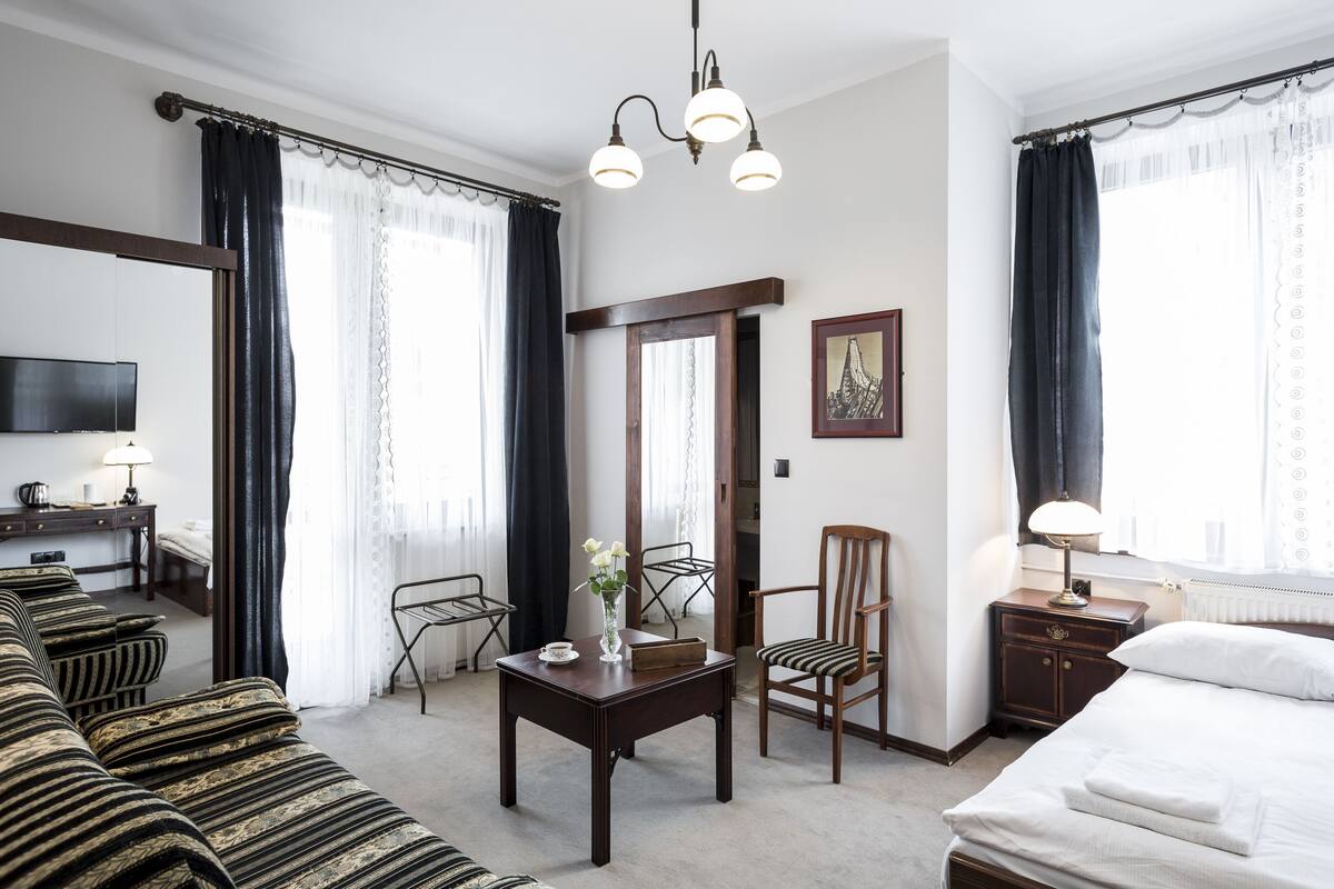 Habitación de hotel con cama, sofá, mesa con un florero, una silla, televisión y ventana con cortinas.