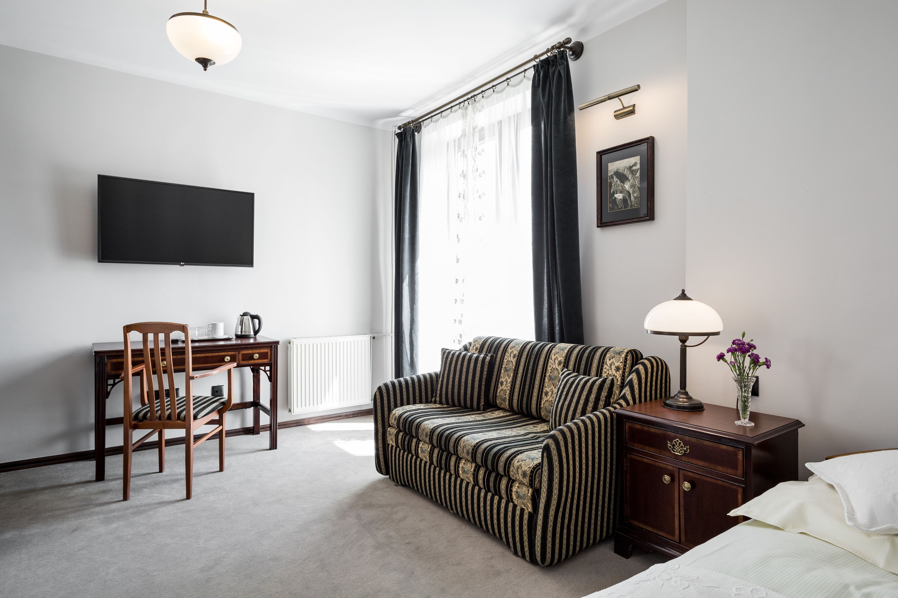 A hotel room with a bed, a sofa, a desk with a chair, a television, a lamp, and a framed picture on the wall.