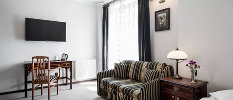 A hotel room with a bed, a sofa, a desk with a chair, a television, a lamp, and a framed picture on the wall.