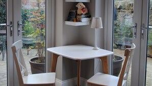 A corner room with a white table and two chairs, a bookshelf with books and decorative items, and a view through glass doors to an outdoor garden. - The Riverhouse (Cardiff)