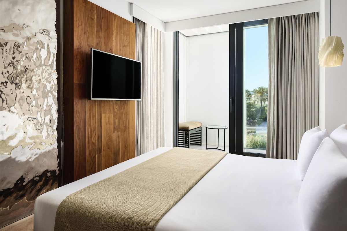 A hotel room with a large bed, a flat-screen TV mounted on a wooden panel, a small round table, and a view of the outdoors through large windows.