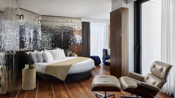 A modern bedroom with a curved bed, a leather armchair, and a large window with curtains.