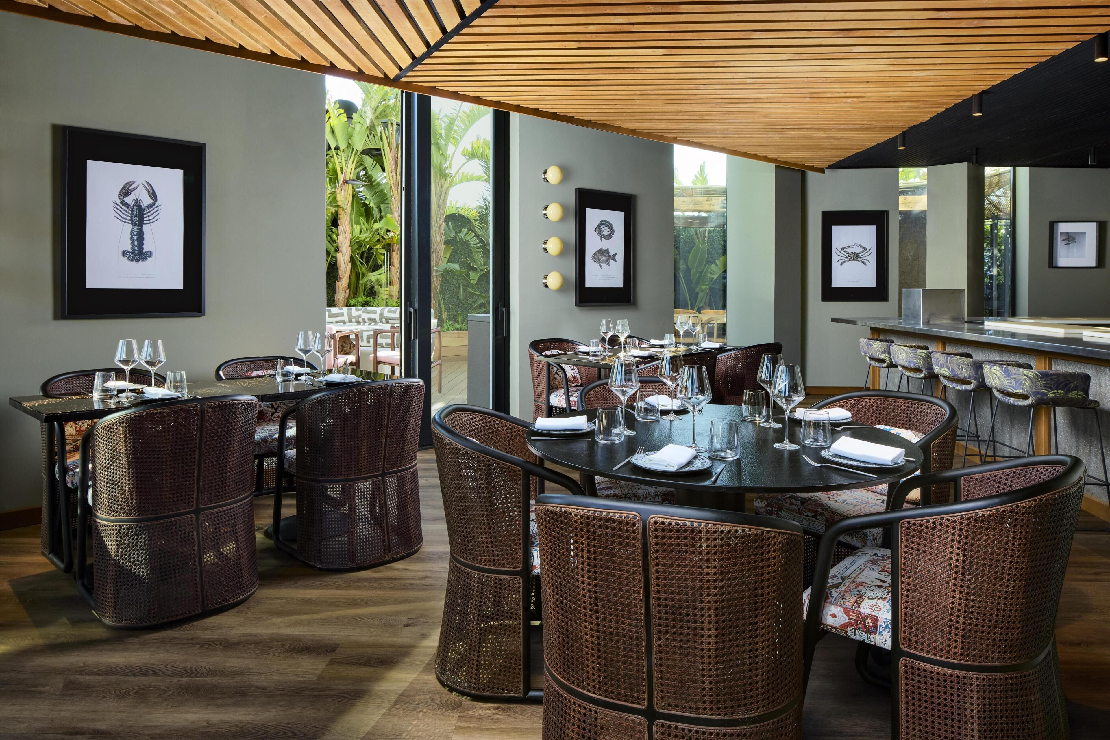 A restaurant with wicker furniture, framed artwork on the walls, and a view of greenery outside.
