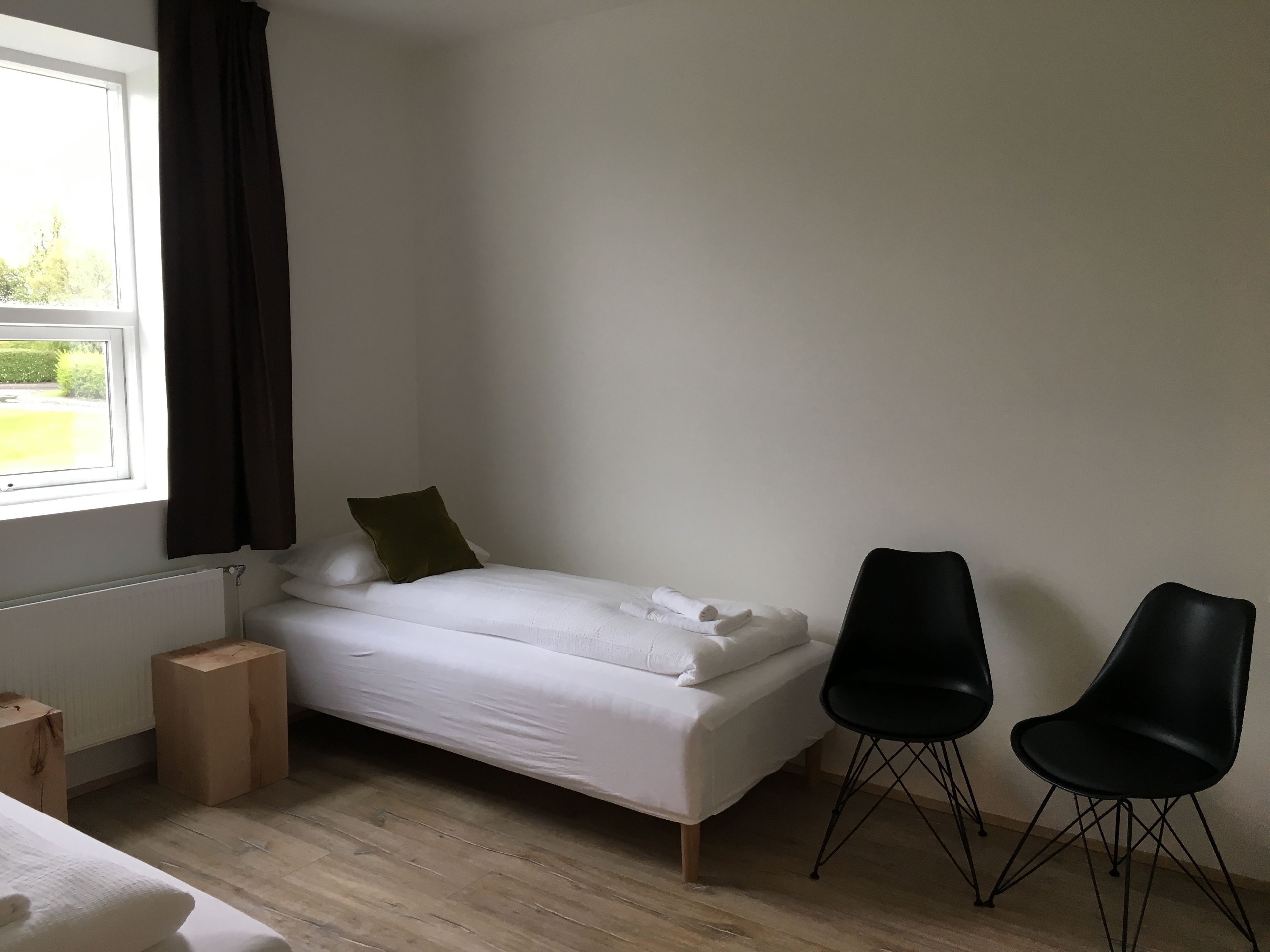 A small, clean room with a single bed, two black chairs, a window with a view of greenery, and a radiator.