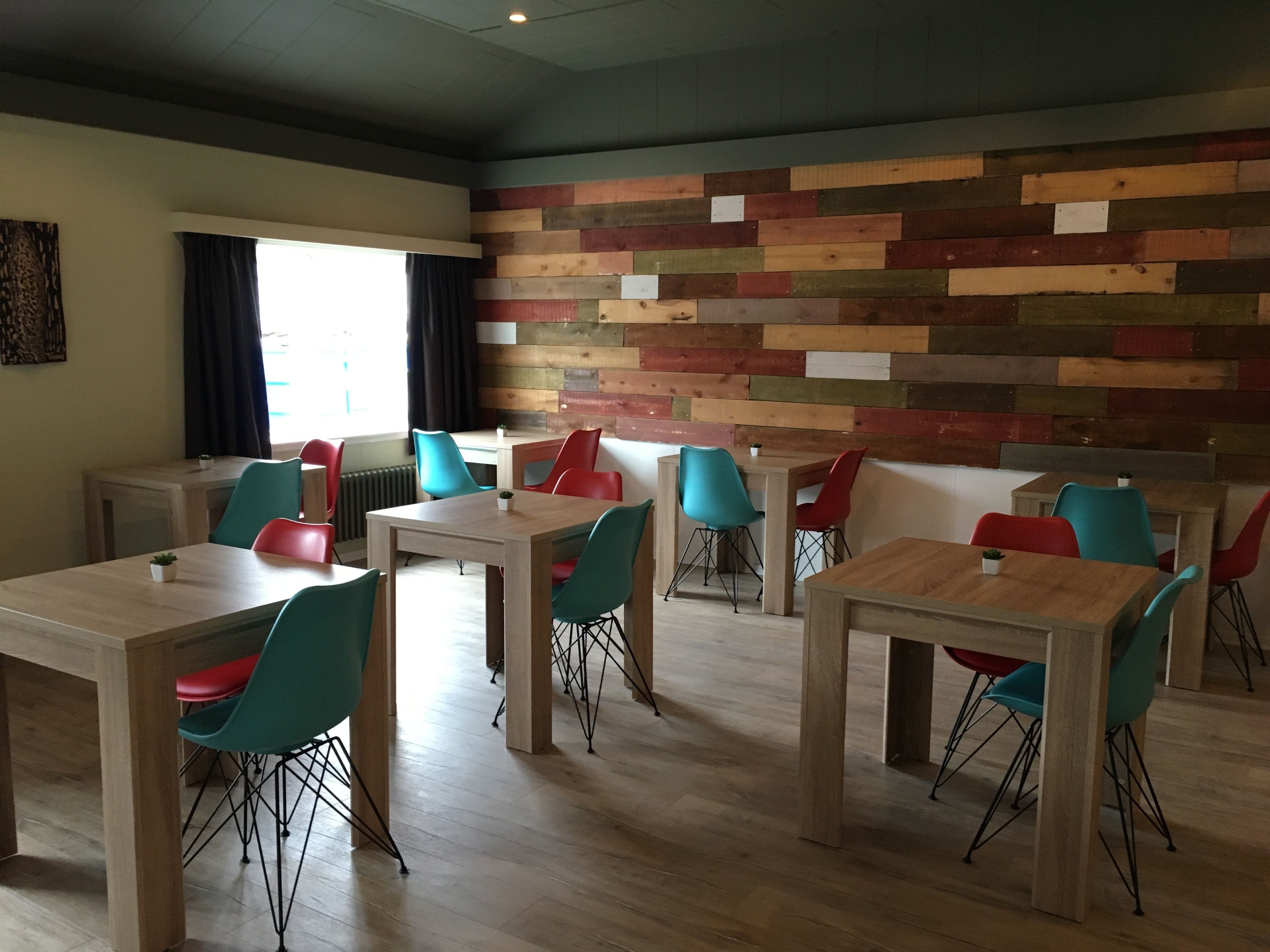 A dining area with wooden tables and chairs, a colorful wooden wall, and a window with curtains.
