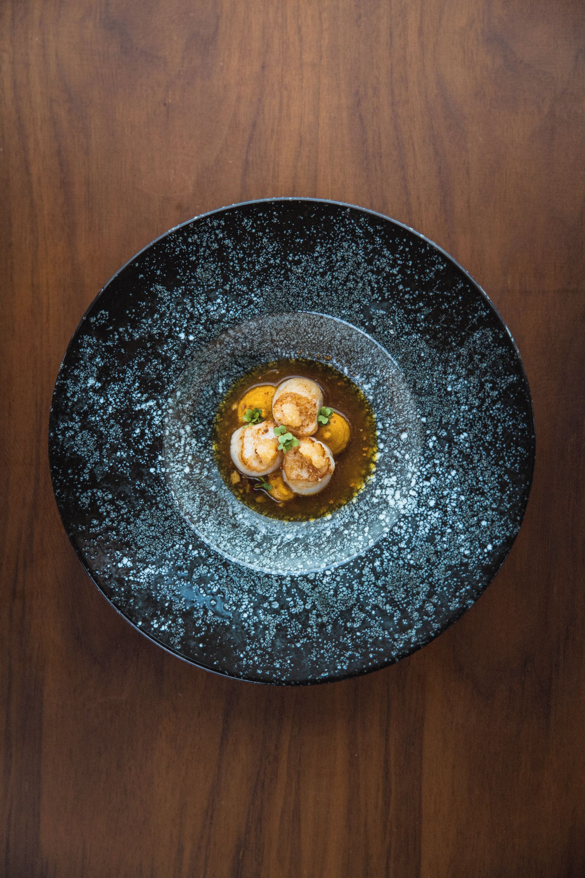 A round black plate with a speckled design, containing three scallops and a garnish, on a wooden surface.