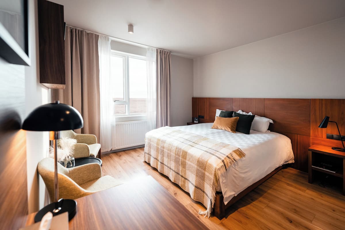 A modern hotel room with a large bed, wooden flooring, a desk, and a chair.