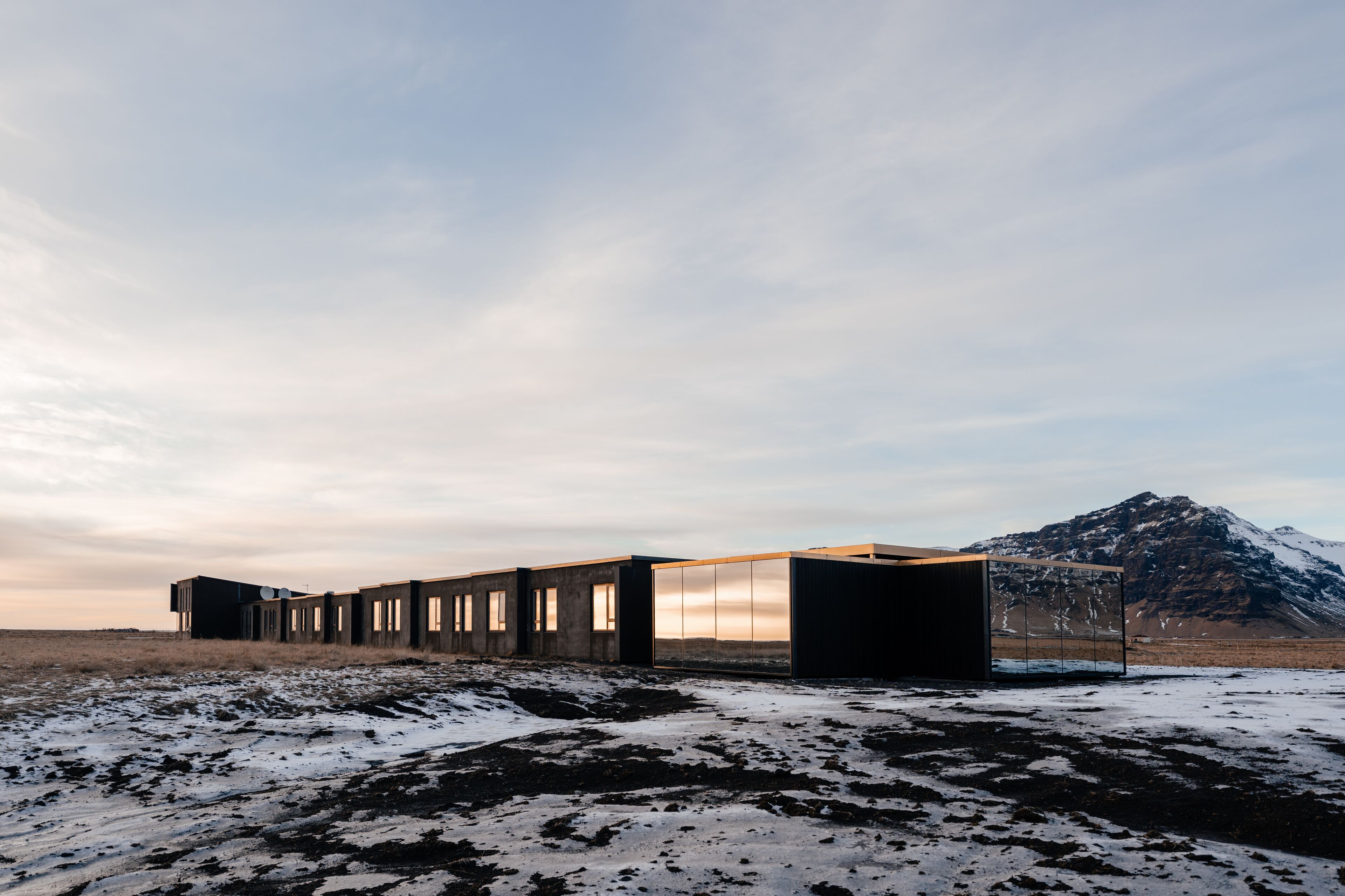 Un edificio moderno con diseño minimalista, que presenta una serie de ventanas rectangulares y un techo plano, ubicado en un paisaje nevado con una montaña al fondo.