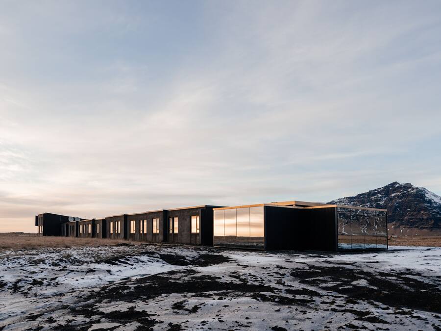 Un edificio moderno con diseño minimalista, que presenta una serie de ventanas rectangulares y un techo plano, ubicado en un paisaje nevado con una montaña al fondo.