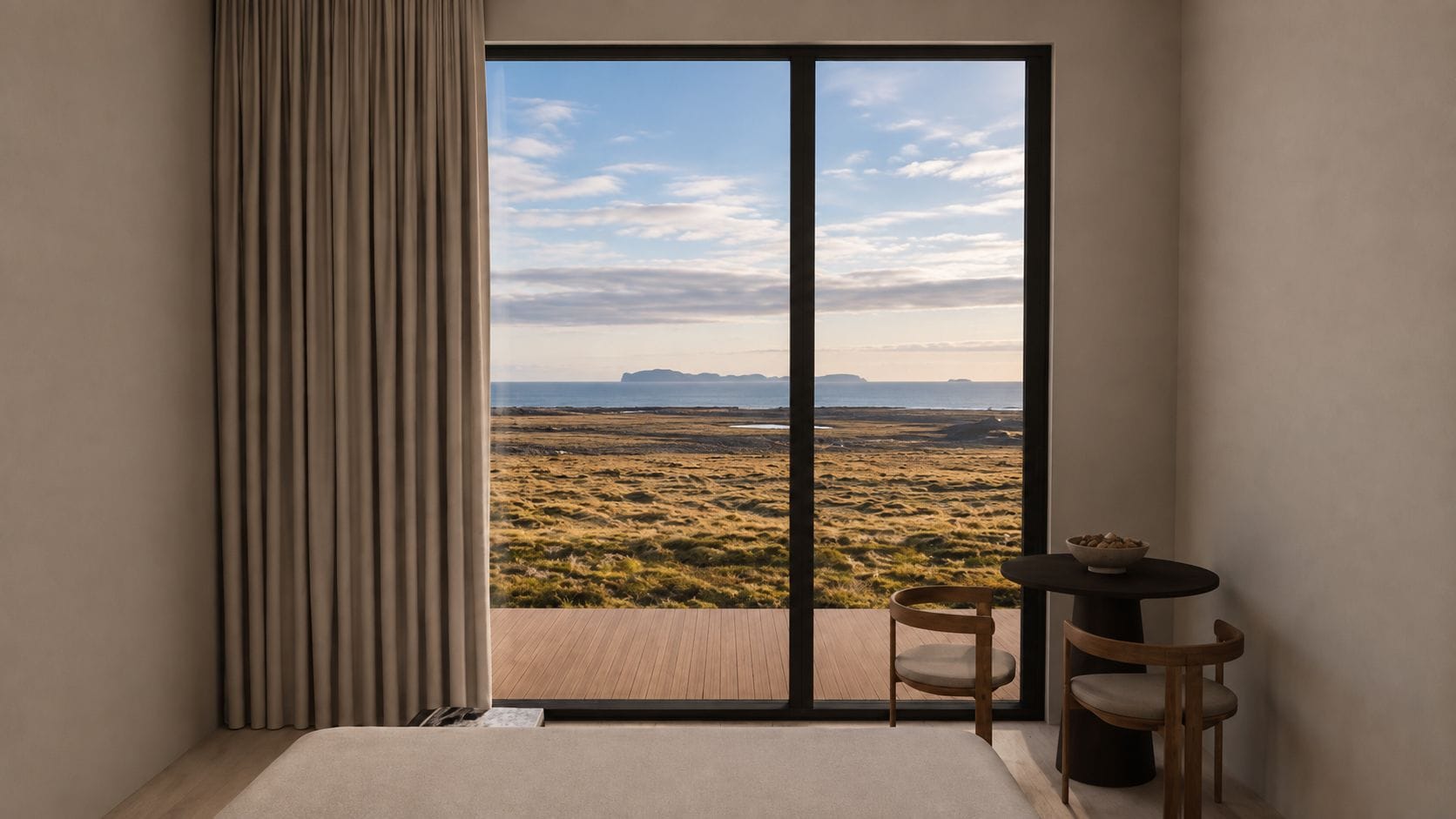 A room with a large sliding glass door leading to a view of a grassy landscape and distant mountains. There is a small round table with a bowl of fruit and two chairs.