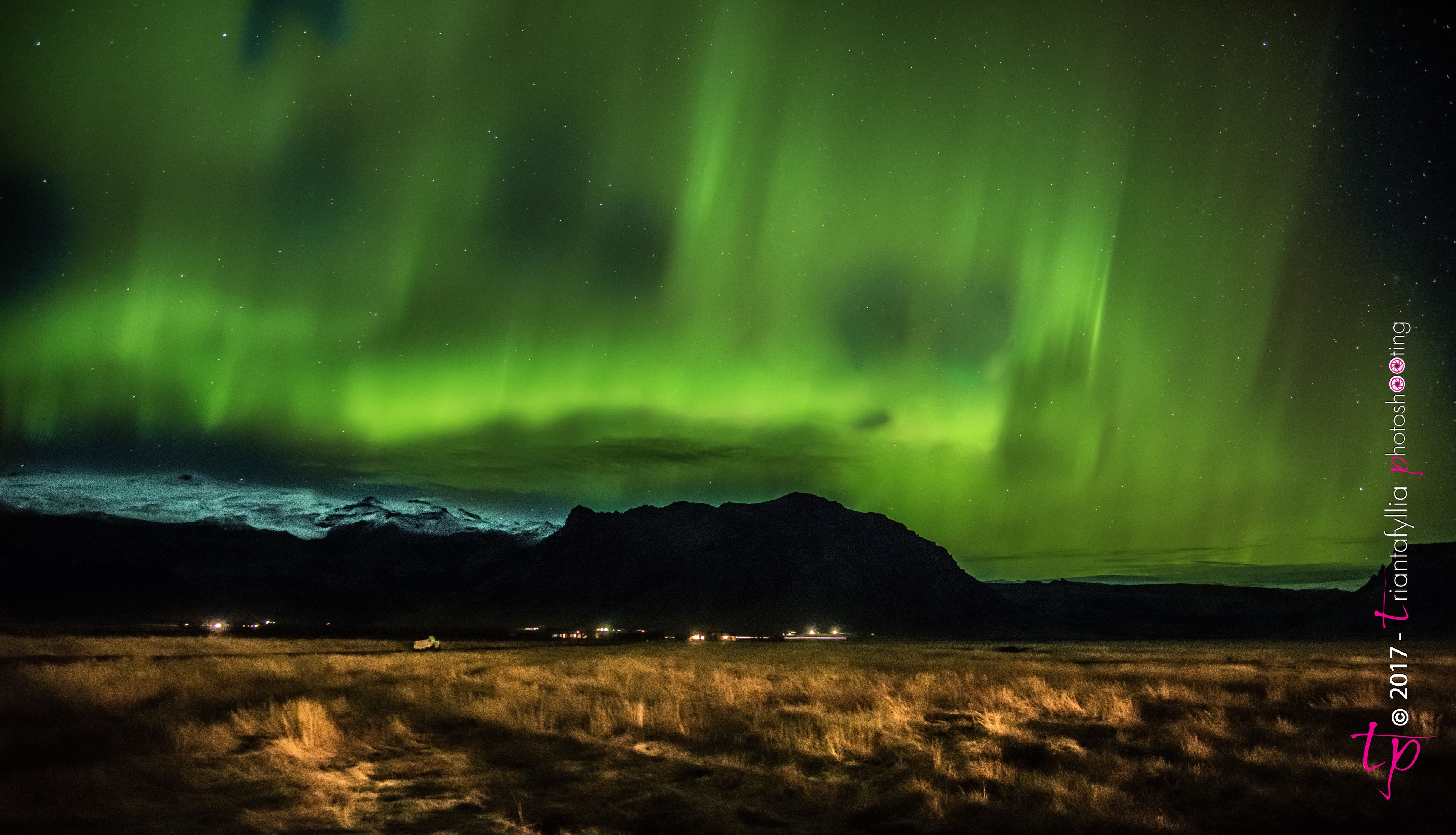 A night sky with the Northern Lights over a mountainous landscape with distant lights.