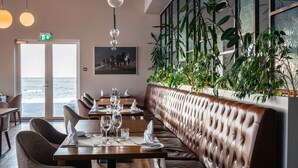 A restaurant with a long wooden bench, tables with chairs, hanging pendant lights, and large windows with a view of the ocean. - UMI Hotel (Rangárþing eystra)