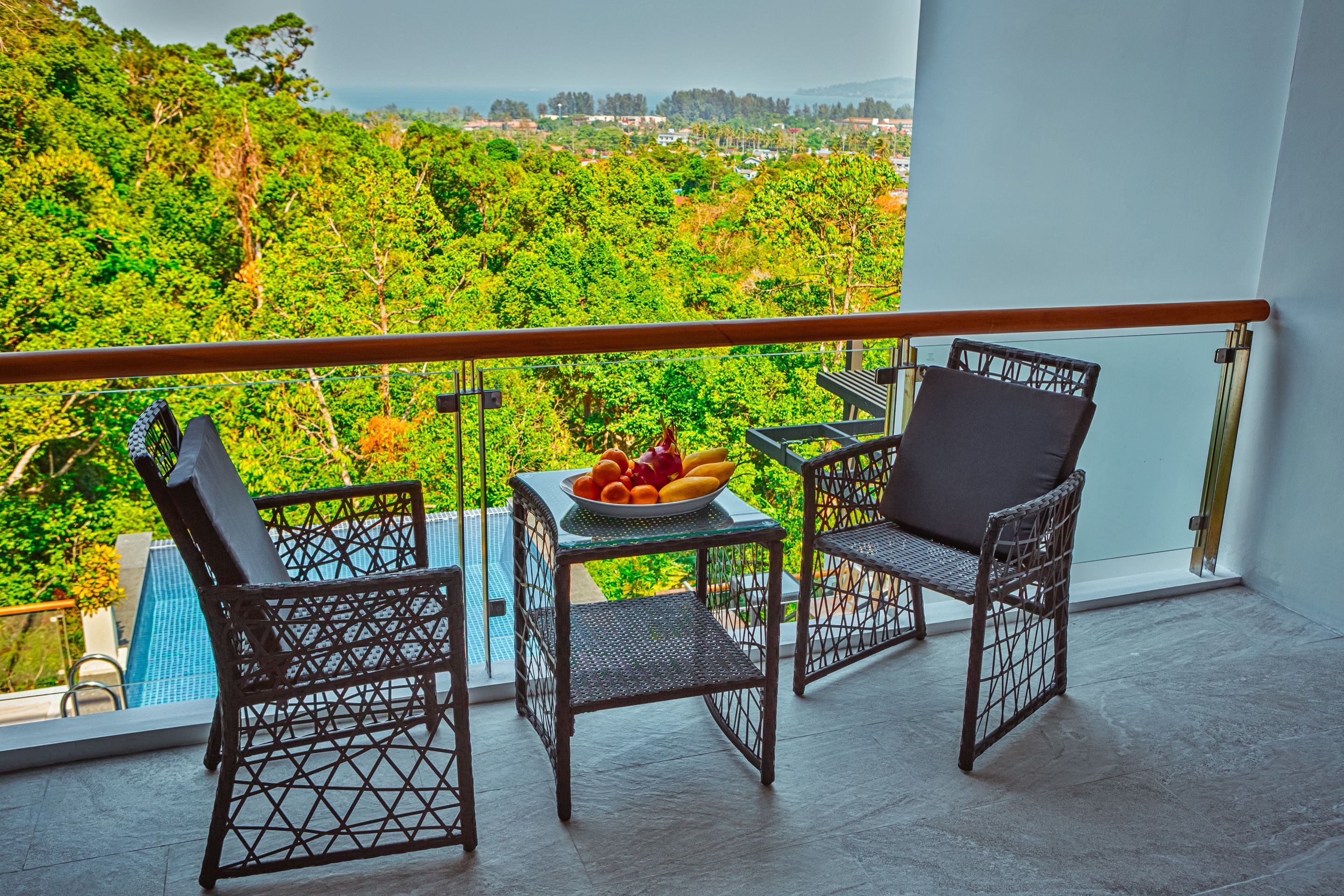 A balcony with wicker chairs and a table with fruit, overlooking a lush green landscape.