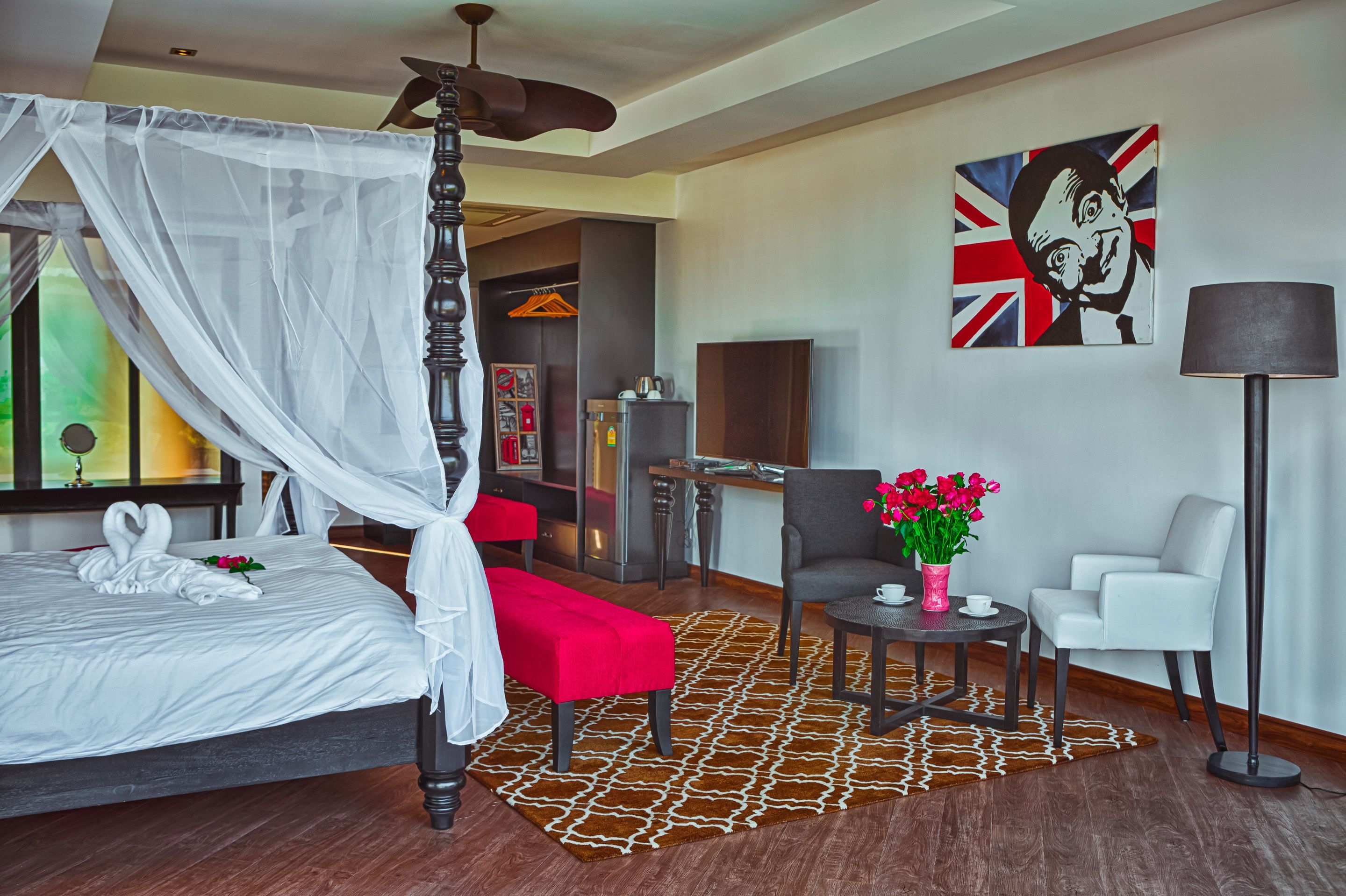 Een slaapkamer met een hemelbed, een rode poef, een kleine tafel met een bloevas, een stoel en een schilderij van de Britse vlag.