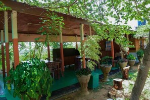 An outdoor covered patio area with tables and chairs, surrounded by potted plants and trees.
