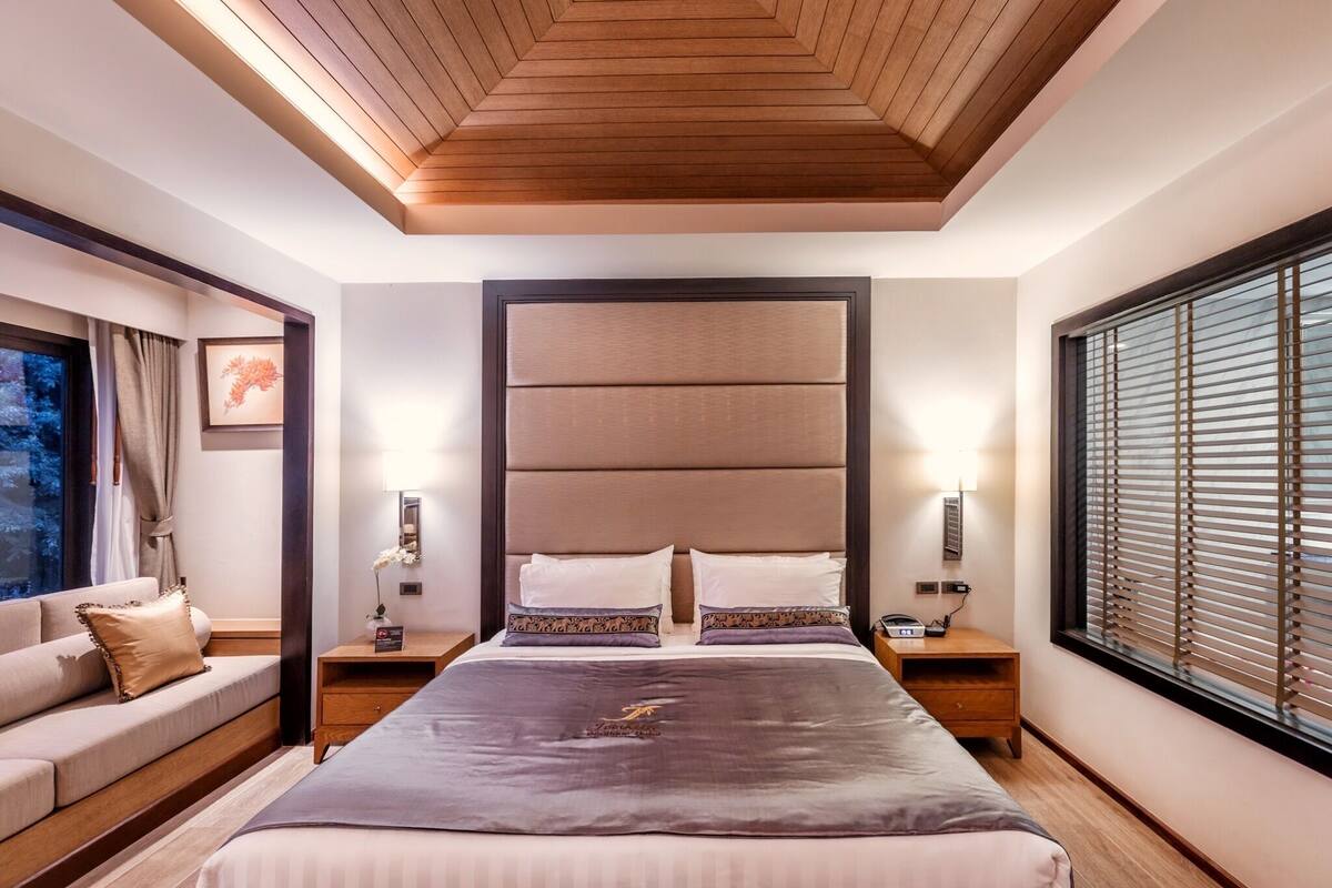 Una habitación de hotel moderna con una cama grande, un sofá, una mesita de noche con lámpara y una ventana con persianas.