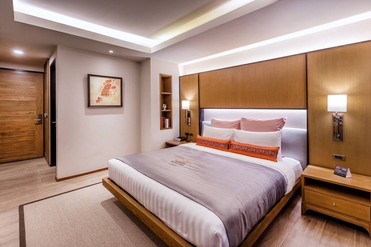Un moderno cuarto de hotel con una cama grande, cabecera de madera, mesitas de noche, un aparador y un cuadro enmarcado en la pared, además de un armario de madera.