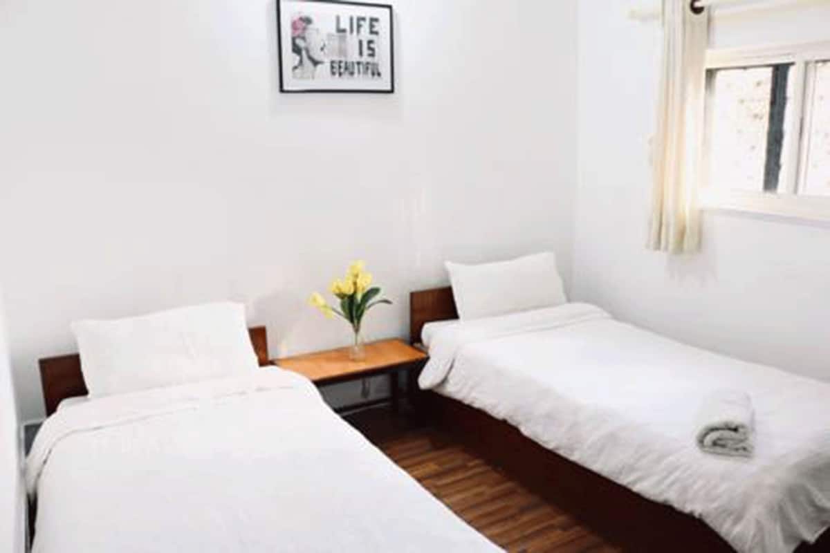 Ein Hotelzimmer mit zwei Einzelbetten, Holzboden, einem kleinen Nachttisch mit Blumenvase und einem Fenster mit Vorhängen.