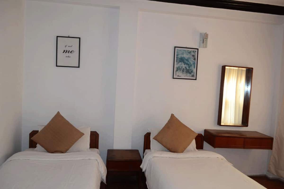Ein Hotelzimmer mit zwei Einzelbetten, einem kleinen Holfnachtkästchen, einem Spiegel und zwei gerahmten Bildern an der Wand.