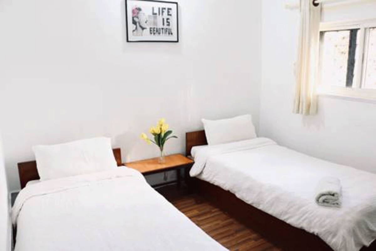 Ein Hotelzimmer mit zwei Einzelbetten, einem kleinen Nachttisch mit Blumenvase, einem gerahmten Motivationsspruch an der Wand und einem Fenster mit Vorhängen.