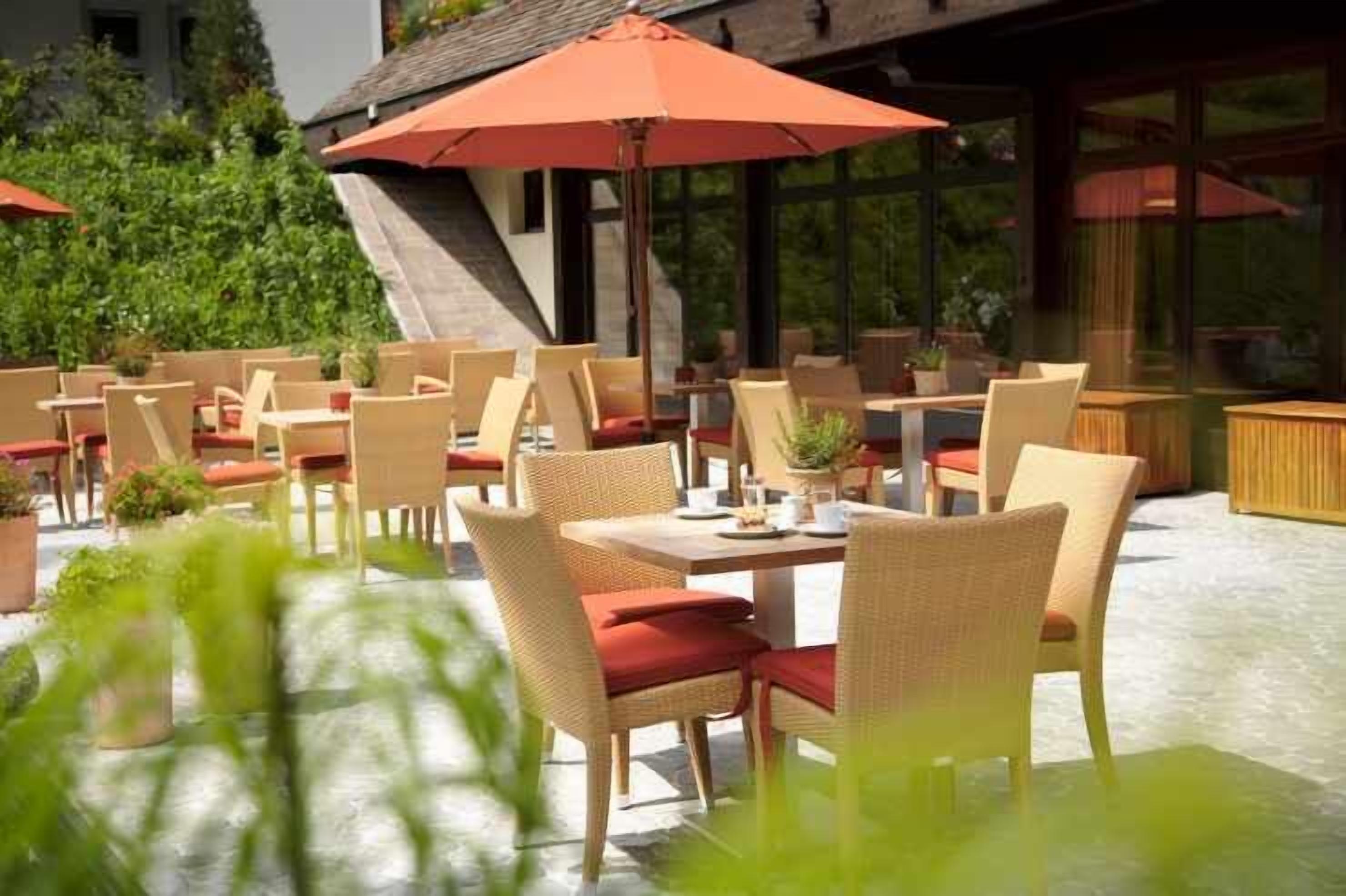 An outdoor seating area with wicker chairs and red cushions, tables with umbrellas, and potted plants.