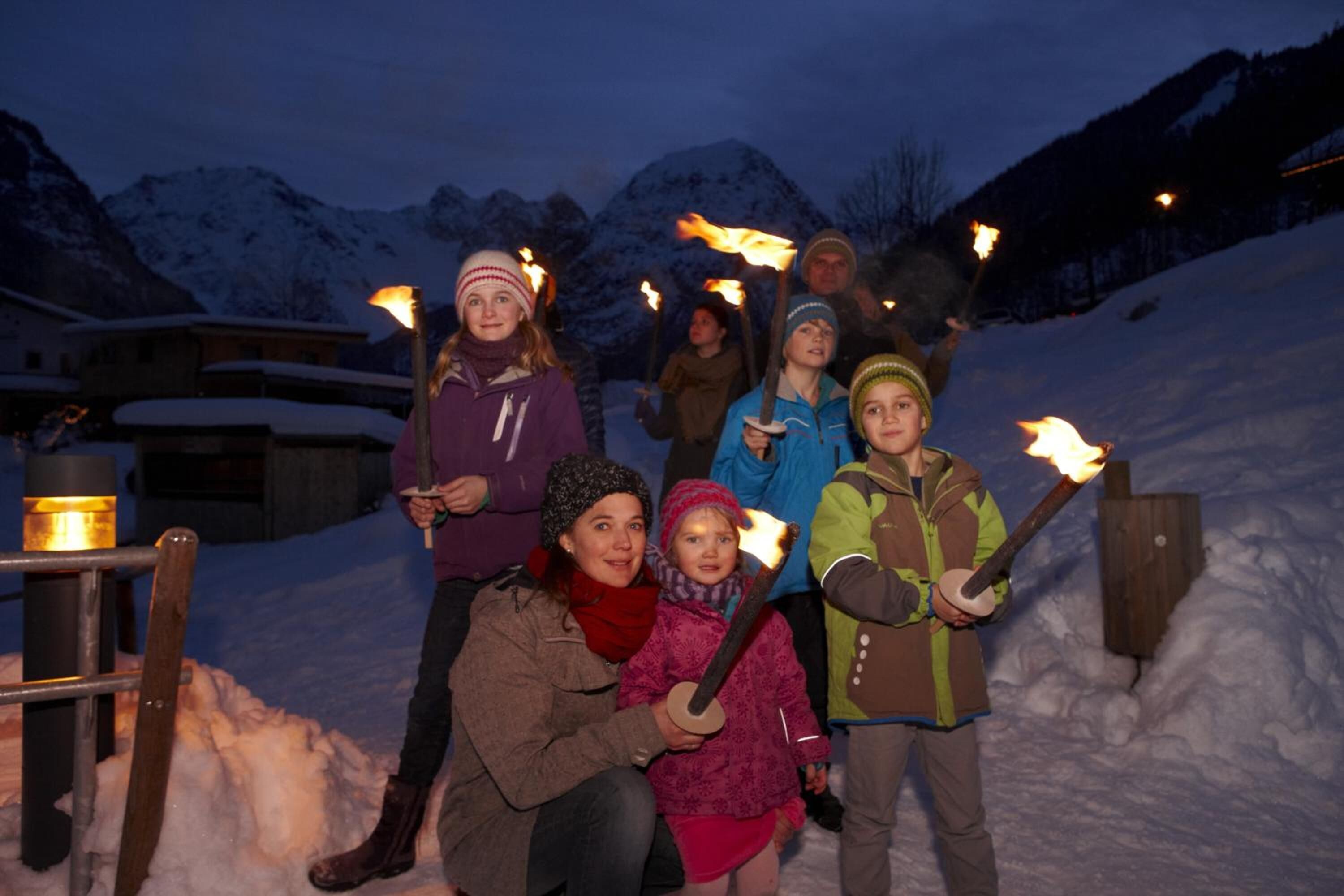 Eine Gruppe von Menschen hält Fackeln in einer verschneiten Winterlandschaft, im Hintergrund Berge.