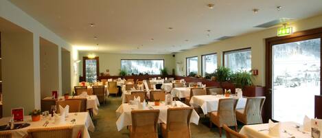 A restaurant with multiple tables and chairs, large windows, and indoor plants.