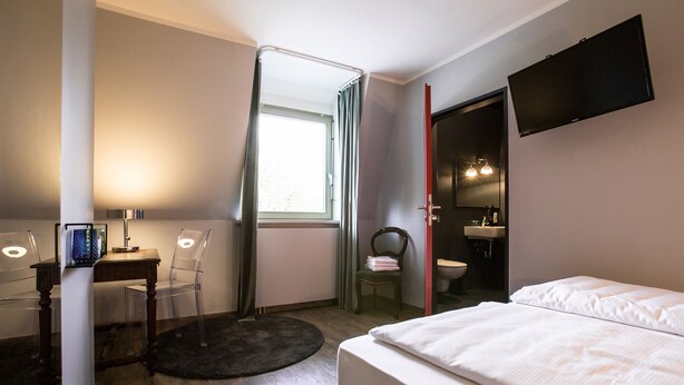 Une chambre d’hôtel comprenant un lit, un bureau avec un ordinateur, une chaise, une fenêtre avec des rideaux, un téléviseur fixé au mur et une salle de bain visible à travers une porte ouverte.