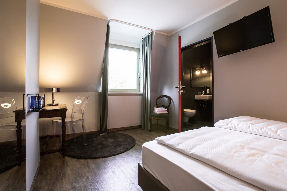 Ein Hotelzimmer mit Bett, Schreibtisch mit Computer, Stuhl, einem Fenster mit Vorhängen, einem an der Wand befestigten Fernseher und einem durchgegangenen Badezimmer.