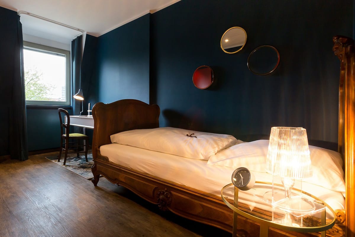 Ein Schlafzimmer mit einem hölzernen Bett, einem Schreibtisch und einem Stuhl. Es gibt ein Fenster mit Vorhängen, eine Lampe und eine Uhr auf einem Nachttisch.