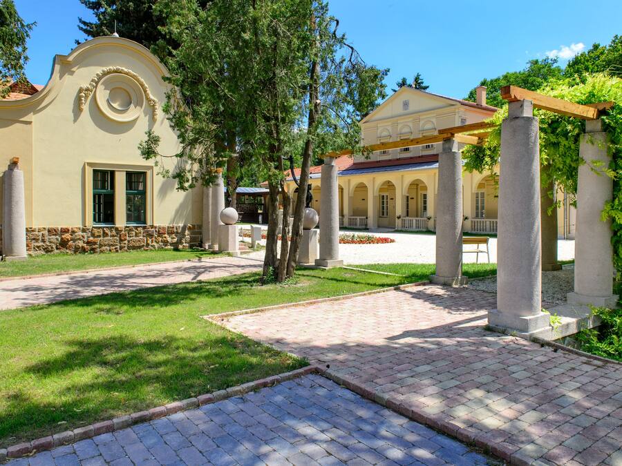 A courtyard with a paved walkway, stone columns, and a building with a yellow facade and arched windows.